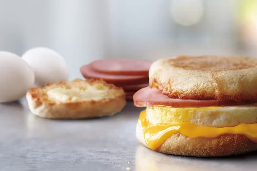 A freshly cracked, 100% cage-free egg on a McDonald’s Egg McMuffin with Canadian-style bacon and melting cheese, beside two whole eggs and a toasted English muffin