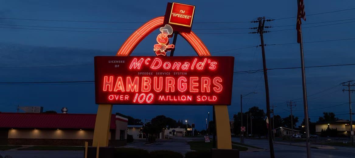 A glowing vintage McDonald's neon sign at dusk — the Speedee chef mascot above “McDonald’s Hamburgers, Over 100 Million Sold”